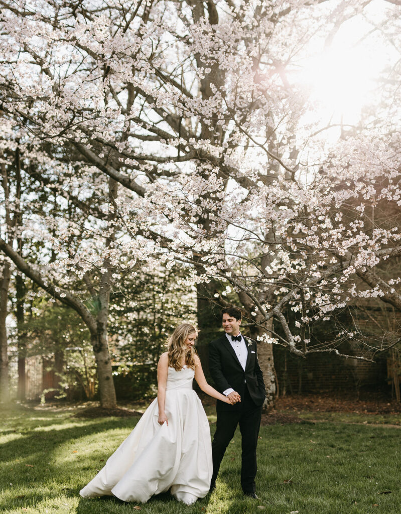 Bride and groom holding hands in the gardens at the Virginia House in Richmond, Virginia, surrounded by blooming trees cherry blossom trees.