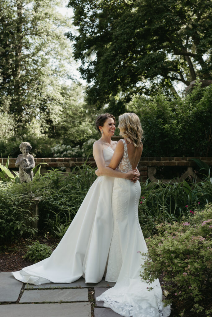 Two brides in wedding gowns hugging in the gardens at the Virginia House in Richmond, Virginia, surrounded by blooming trees and historic estate architecture.
