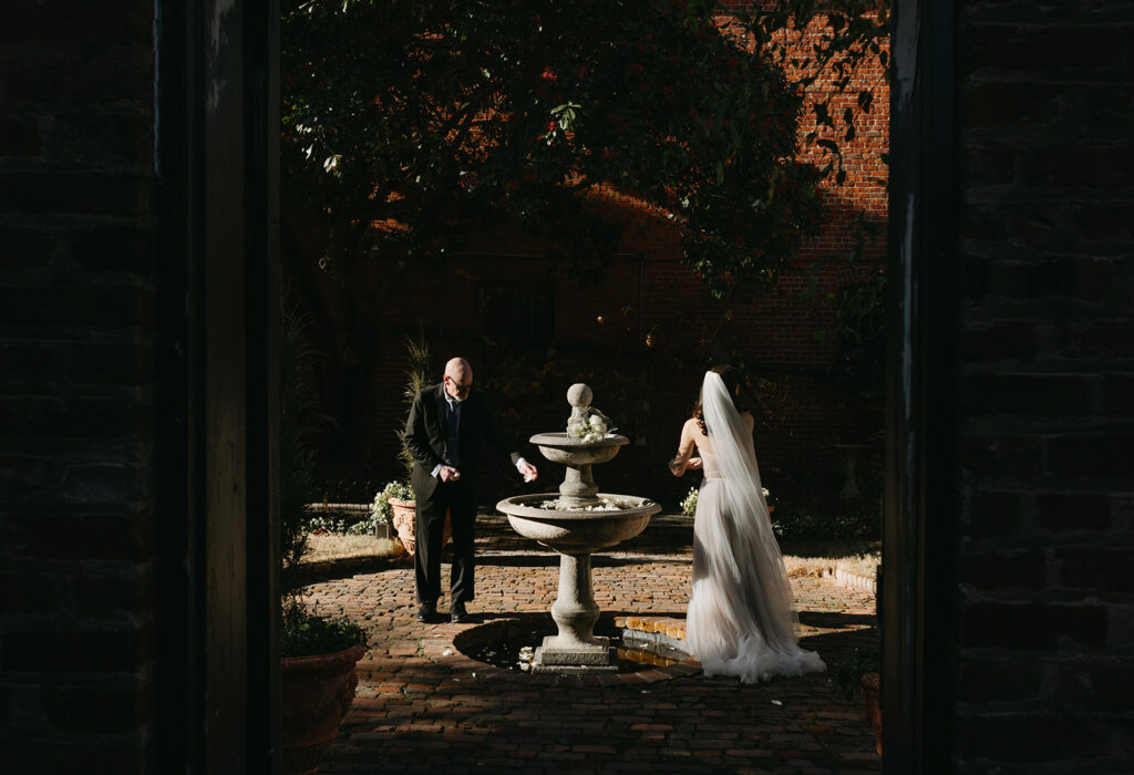 Richmond winter micro wedding at the Poe museum. Image of Bride and dad placing flower petals in the courtyard fountain.