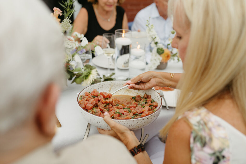Reception rooftop at Summers Roof and Cellar, decorated with summer florals and family-style tables. Guests dining and passing plates.