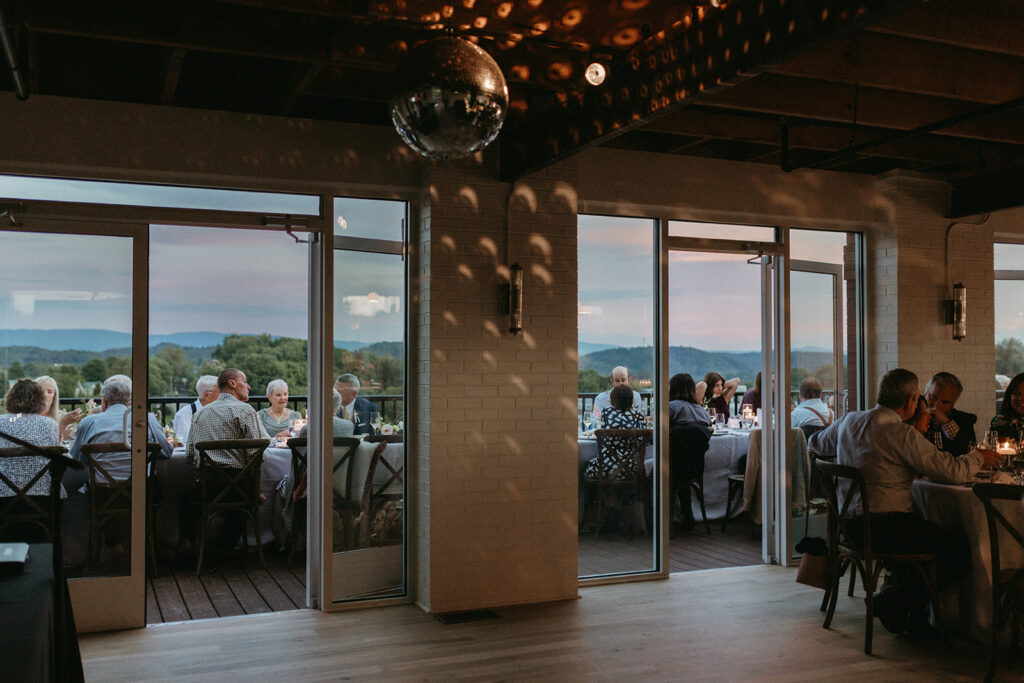 Reception rooftop at Summers Roof and Cellar, decorated with summer florals and family-style tables. Guests dining and passing plates.