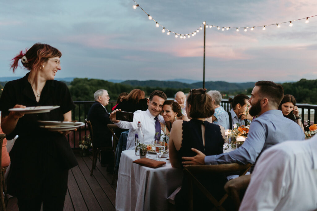 Reception rooftop at Summers Roof and Cellar, decorated with summer florals and family-style tables. Guests dining and passing plates.