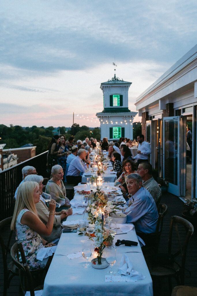 Reception rooftop at Summers Roof and Cellar, decorated with summer florals and family-style tables. Guests dining and passing plates.